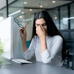 Frau reibt sich die Augen, hält Brille, Computerbildschirm und Tastatur im Büro. Sie braucht eine Pause nach der 20-20-20 Regel.