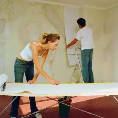 Ein Paar tapeziert eine gestrichene Wand. Die Frau misst Tapete auf einem Tisch, während der Mann sie an der Wand anbringt.