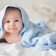 Ein lächelndes Baby liegt auf einem Bett, eingewickelt in ein hellblaues Handtuch. Die Frage "wie viel Babykleidung braucht man" beschäftigt viele Eltern.