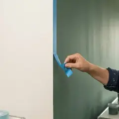 Frau klebt Klebeband an die Wand, um eine klare Trennlinie zu schaffen. Sie wird die Wand nicht bis zur Decke streichen, sondern nur einen Teil davon.