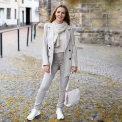 Frau in beige grau farbe Outfit mit Mantel, Schal, Hose und Tasche steht auf Kopfsteinpflaster.