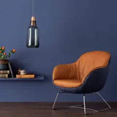 Ein Sessel mit cognacfarbenem Leder und dunkelblauen Akzenten steht vor einer Wand in tiefen blau nuancen. Daneben ein Regal mit Büchern und einer Vase.