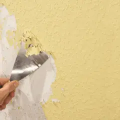 Hand schabt mit Spachtel alte Tapetenreste von gelber Wand.