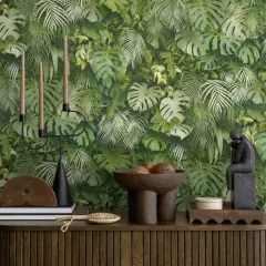 Dschungel-Tapete mit Monstera und Farnen. Ein Kerzenständer, Bücher und eine Skulptur stehen auf einem Holzmöbel. Der Unterschied Vlies und Papiertapete wird hier nicht gezeigt.