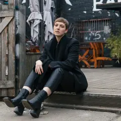 Frau in schwarzem Outfit sitzt vor Holztür. Sie überlegt, wieviel spart man wenn man keine kleidung mehr kaufen muss.