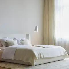 Ein helles Schlafzimmer mit einem großen Bett, das mit weißer Bettwäsche und einer cremefarbenen Decke bedeckt ist. Ein moderner Teppich bedeckt das ganze Zimmer und verleiht ihm Gemütlichkeit.