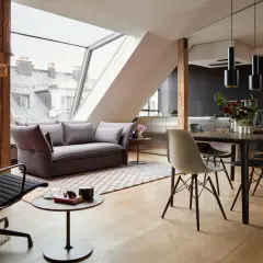 Modernes Wohnzimmer mit Dachschräge, wo sich Sofa und Esstisch kombinieren. Holzbalken und große Fenster schaffen ein luftiges Ambiente.