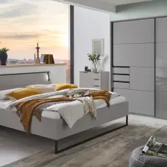 Modernes Schlafzimmer mit grauem Bett, Nachttischen und einem großen Kleiderschrank. Ein Fenster gibt den Blick auf eine Stadt bei Sonnenuntergang frei.