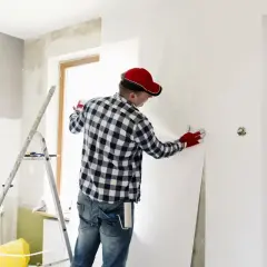Mann mit roter Kappe tapeziert Wand. Verschiedene Raufasertapeten Arten sind für Renovierungen beliebt.