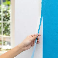 Hand zieht blauen Malerkrepp von weißer Wand ab. Das ist ein wichtiger Schritt, wenn man wissen will, was man zum Streichen braucht.