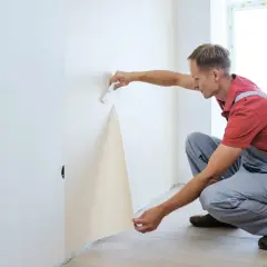 Mann kleistert Vliestapete an die Wand. Er hält ein Stück Tapete und streicht es mit einem Werkzeug glatt.
