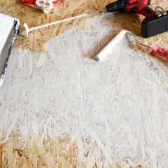 OSB-Platten streichen: Ein weißer Farbroller und eine Farbwanne liegen bereit. Ein Akkuschrauber und Handschuhe deuten auf ein DIY-Projekt hin.