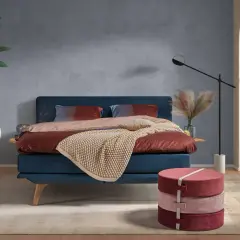 Ein blaues Bett mit rustikaler, grauer Wandfarbe im Hintergrund. Dekorative Kissen und eine warme Decke laden zum Entspannen ein.