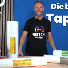 Mann vergleicht Malervlies vs Rauhfaser Kosten. Mehrere Rollen Tapeten liegen auf einem Tisch, ein Plüschtier schaut hervor.