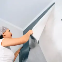 Frau mit Kopftuch entfernt Tapete von Decke. Sie zieht das graue Material ab, um die weiße Wand darunter freizulegen.