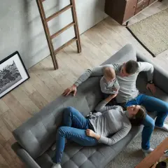 Familie entspannt auf Sofa, ein Baby auf dem Arm. Minimalismus Lebensstil in einem aufgeräumten, hellen Wohnzimmer mit Holzboden.