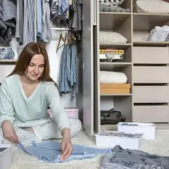 Frau sortiert Kleidung vor einem offenen Kleiderschrank. Sie fragt sich, wo sie mit dem **Haus ausmisten wo anfangen** soll.