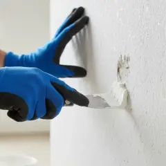 Mit blauen Handschuhen wird mit einer Spachtelmasse eine beschädigte Stelle an einer Raufasertapete ausgebessert.
