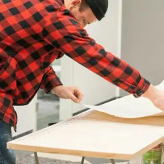Mann in kariertem Hemd bereitet sich auf tapezieren ohne Kleister vor. Er zieht die Schutzfolie von einer selbstklebenden Tapete ab.