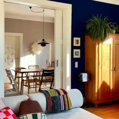 Ein Wohnzimmer mit einer tiefblauen Wand, einem Holzkasten, Pflanzen und einem gemütlichen Sofa. Durch eine Tür ist ein Esszimmer mit Holztisch und Stühlen zu sehen.