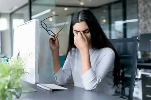 Frau reibt sich die Augen, hält Brille, Computerbildschirm und Tastatur im Büro. Sie braucht eine Pause nach der 20-20-20 Regel.