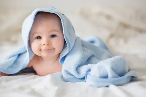 Ein lächelndes Baby liegt auf einem Bett, eingewickelt in ein hellblaues Handtuch. Die Frage "wie viel Babykleidung braucht man" beschäftigt viele Eltern.
