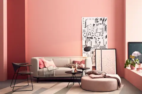 Gemütliches Wohnzimmer mit rosa Wand, bequemer Couch, Kissen und einem Pouf. Ein moderner Sessel und Kunstwerke ergänzen den Raum.