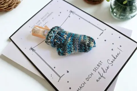 Gestrickte Socke mit Geld als Geschenk. Die Karte fragt "wie viele Socken braucht man?" und ermutigt, sich selbst auf die Socken zu machen.