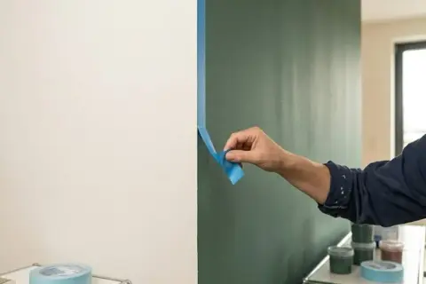 Frau klebt Klebeband an die Wand, um eine klare Trennlinie zu schaffen. Sie wird die Wand nicht bis zur Decke streichen, sondern nur einen Teil davon.