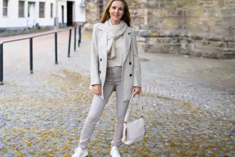 Frau in beige grau farbe Outfit mit Mantel, Schal, Hose und Tasche steht auf Kopfsteinpflaster.