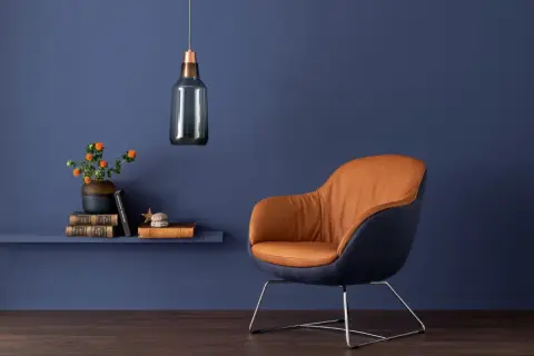Ein Sessel mit cognacfarbenem Leder und dunkelblauen Akzenten steht vor einer Wand in tiefen blau nuancen. Daneben ein Regal mit Büchern und einer Vase.