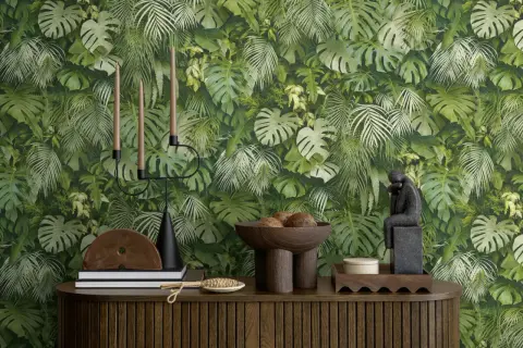 Dschungel-Tapete mit Monstera und Farnen. Ein Kerzenständer, Bücher und eine Skulptur stehen auf einem Holzmöbel. Der Unterschied Vlies und Papiertapete wird hier nicht gezeigt.