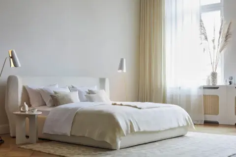 Ein helles Schlafzimmer mit einem großen Bett, das mit weißer Bettwäsche und einer cremefarbenen Decke bedeckt ist. Ein moderner Teppich bedeckt das ganze Zimmer und verleiht ihm Gemütlichkeit.