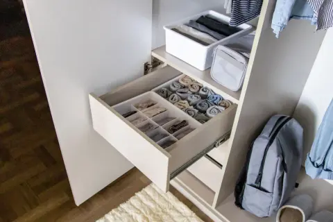 Schrank mit ordentlich gefalteten Kleidungsstücken in Schubladen und Körben. Ein Rucksack und Jeans hängen daneben. Zu viele Klamotten zu wenig Platz? Hier ist die Lösung!