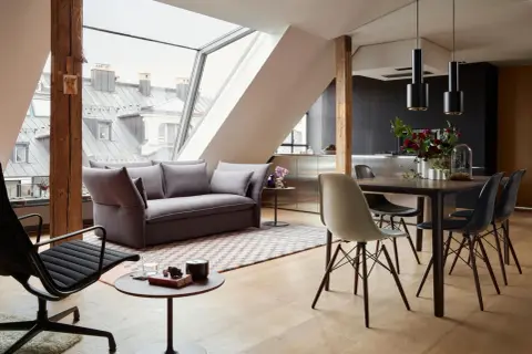Modernes Wohnzimmer mit Dachschräge, wo sich Sofa und Esstisch kombinieren. Holzbalken und große Fenster schaffen ein luftiges Ambiente.