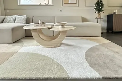 Ein Wohnzimmer mit einem beigen Ecksofa, einem runden Couchtisch und einem modernen Teppich im geometrischen Design. Dieser Teppich-Trend bringt Ruhe und Stil in jeden Raum.
