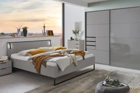 Modernes Schlafzimmer mit grauem Bett, Nachttischen und einem großen Kleiderschrank. Ein Fenster gibt den Blick auf eine Stadt bei Sonnenuntergang frei.