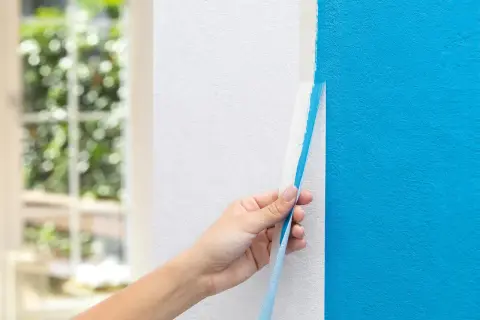 Hand zieht blauen Malerkrepp von weißer Wand ab. Das ist ein wichtiger Schritt, wenn man wissen will, was man zum Streichen braucht.