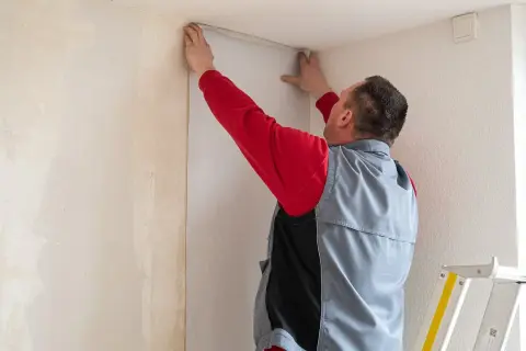 Mann tapeziert eine Wand in einer Wohnung. Er hält eine Tapetenbahn an die Decke, um sie anzubringen.