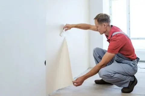 Mann kleistert Vliestapete an die Wand. Er hält ein Stück Tapete und streicht es mit einem Werkzeug glatt.