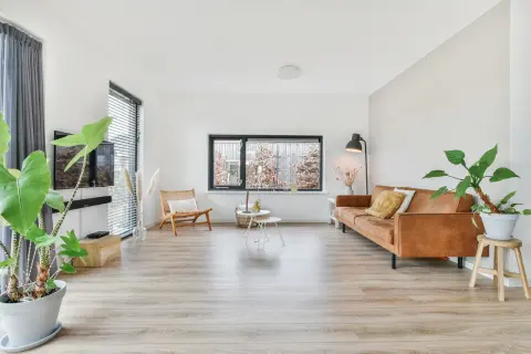 Ein heller Boden im Wohnzimmer reflektiert das Licht. Ein braunes Ledersofa, ein Korbstuhl und Pflanzen schaffen eine gemütliche Atmosphäre.