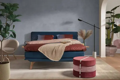 Ein blaues Bett mit rustikaler, grauer Wandfarbe im Hintergrund. Dekorative Kissen und eine warme Decke laden zum Entspannen ein.