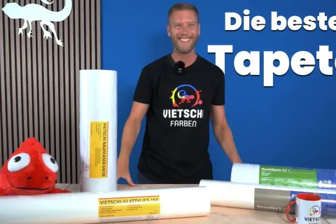 Mann vergleicht Malervlies vs Rauhfaser Kosten. Mehrere Rollen Tapeten liegen auf einem Tisch, ein Plüschtier schaut hervor.