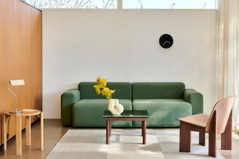 Ein Wohnzimmer im **Scandi Style** mit einem grünen Sofa, einem Holzsessel, einem Beistelltisch mit Lampe und einem Couchtisch mit gelben Blumen.