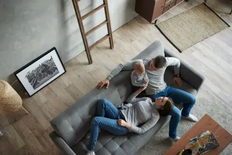 Familie entspannt auf Sofa, ein Baby auf dem Arm. Minimalismus Lebensstil in einem aufgeräumten, hellen Wohnzimmer mit Holzboden.