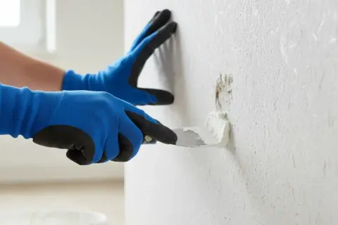 Mit blauen Handschuhen wird mit einer Spachtelmasse eine beschädigte Stelle an einer Raufasertapete ausgebessert.