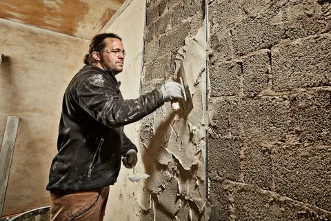 Mann verputzt eine grobe Ziegelwand mit einer Kelle. Der Putz wird gleichmäßig aufgetragen, um die Oberfläche zu glätten.