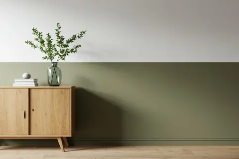 Ein Sideboard aus Holz mit Büchern und einer Vase mit Zweigen steht vor einer zweifarbig gestrichenen Wand. Die untere Hälfte der Wand ist in einem erdigen Grün gehalten, die obere Hälfte in Weiß.