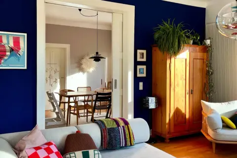 Ein Wohnzimmer mit einer tiefblauen Wand, einem Holzkasten, Pflanzen und einem gemütlichen Sofa. Durch eine Tür ist ein Esszimmer mit Holztisch und Stühlen zu sehen.