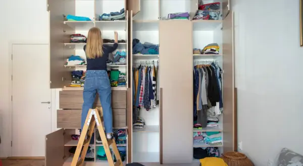Mädchen auf Trittleiter sortiert Kleidung im offenen Kleiderschrank. So miste ich richtig aus: Kleidung wird auf Regalen und Kleiderstangen verstaut.